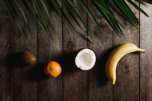 Broken coconut, kiwi, tangerine, orange, banana on brown wooden background with palm leaf. White coconut pulp. High quality photo