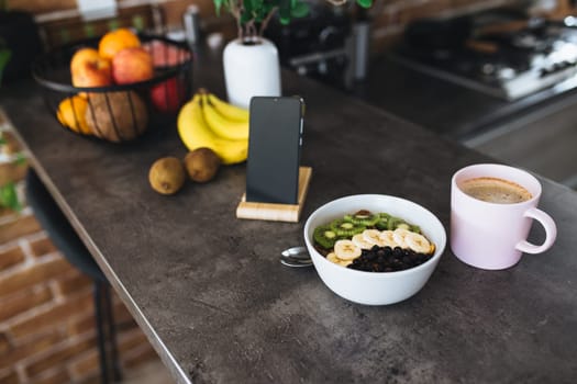 Pink coffee cup, bowl with chopped tropical fruits kiwi and banana, blueberries, spoon and mobile phone on bar counter in stylish loft kitchen. Blurred background. High quality photo
