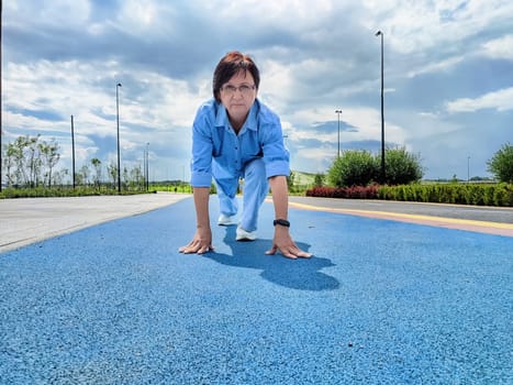 A woman is in a starting position on a vibrant blue track surrounded by greenery. Bright sunlight illuminates the area, creating a lively atmosphere.