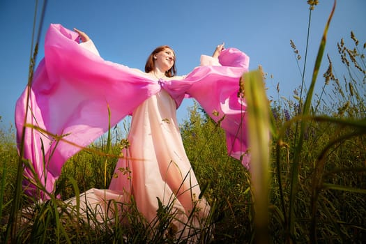 A woman twirls gracefully in a sunlit field, draped in vibrant pink fabric, with green grass swaying around her under a bright sky.