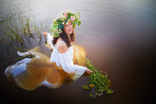 Slavic plump chubby girl in long white dress on the feast of Ivan Kupala with flowers and water in a river or lake on summer evening