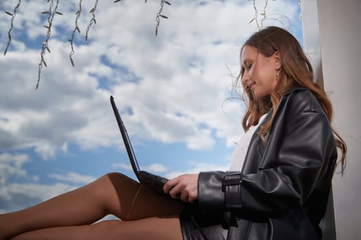A young woman focuses on her laptop while sitting outdoors, enjoying a peaceful moment under a sunny sky.