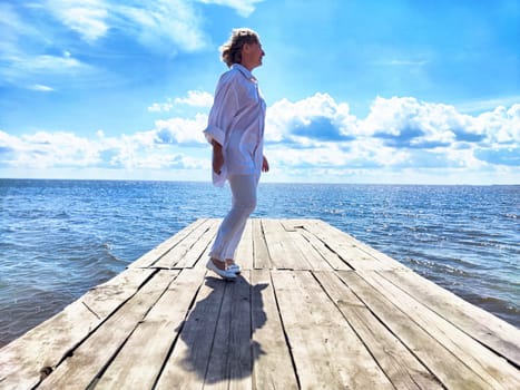 Mature woman stands confidently on a wooden pier, soaking in the beauty of the ocean and sky during her travel adventure.