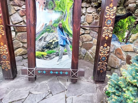 A modern woman takes a funny selfie in a curved mirror while exploring a park filled with greenery and stone features.