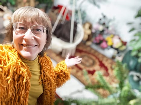 A middle-aged woman with blonde hair presents her vibrant indoor garden filled with greenery and flowers.