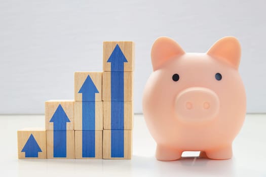 A piggy bank with wood blocks stacking as step stair with blue arrows up. Concept: Successful business, Business growth, Savings growth, Interest rate. Long term savings.