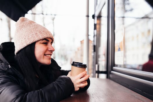 Girl smiling, holds paper cup of coffee in hand near cafe. Blurred background. High quality photo