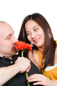 Double head-and-shoulder portrait of a young heterosexual couple looking each other in the eye against a white background with an orange gerbera flower in the foreground.