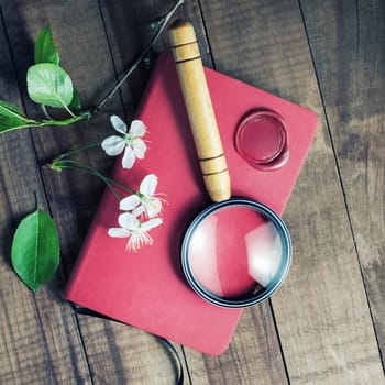 Red notebook, magnifier, wax seal, herry flowers and green leaves on vintage wood table background.