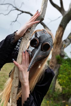 Blonde woman in a crow mask posing near a withered tree in nature, creating an aura of mystery and beauty.