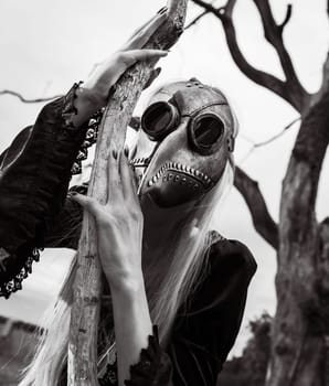 Blonde woman in a crow mask posing near a withered tree in nature, creating an aura of mystery and beauty.