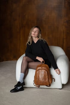 A girl in a warm sweater, a black short skirt, nylon tights, black loafers and gray socks is posing with a brown backpack