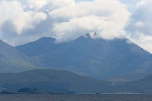 Distant road over the water winds towards a huge rocky mountain on a scottish island. Bright sunlit clouds cover the top of the mountain. Landscape.