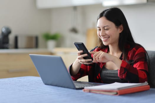 Asian student using phone and laptop in the kitchen at home
