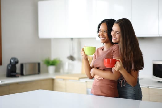 Lesbian interracial couple drinking in a kitchen looking away at home