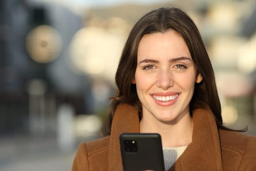 Front view portrait of a happy woman in winter holding phone looking at camera