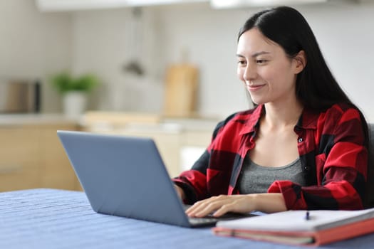 Satisfied asian student writing on laptop in the kitchen at home