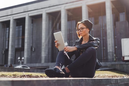 A fashionably dressed hipster girl in a knitted hat uses an electronic tablet sitting outside in the city
