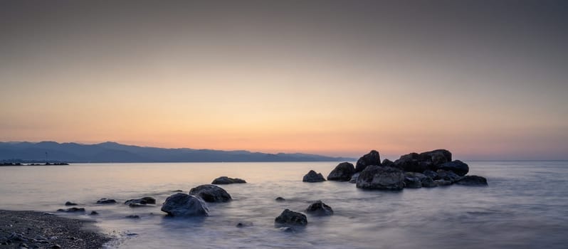 Serene dusk envelops the shoreline, illuminating smooth stones while soft waves lap gently against them.