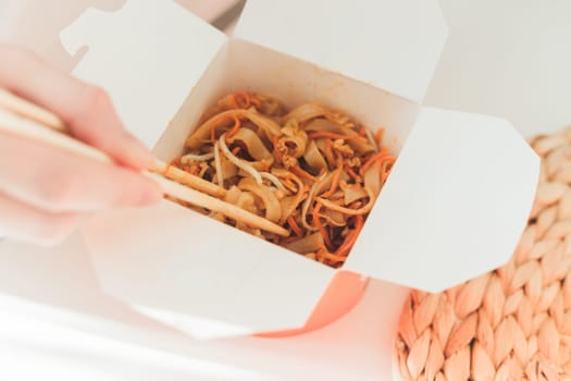 Wok noodles in takeaway box. Woman eating with chopsticks, close up view on female hands. Chinese traditional food with vegetables and seafood.
