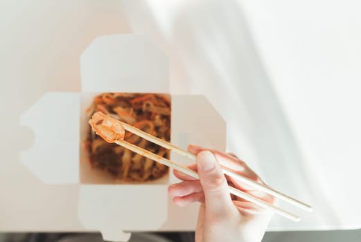 Wok noodles in takeaway box. Woman eating with chopsticks, close up view on female hands. Chinese traditional food with vegetables and seafood.