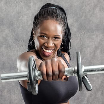 Close up portrait of sportive black teen with braids holding dumbbell in front of face. Young woman in sportswear with vigorous face expression against grey background.