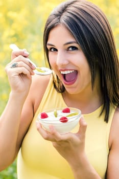 Close up portrait of attractive healthy girl showing yogurt bowl with fruit against yellow flower field.Young woman with open mouth about to eat yogurt on spoon.