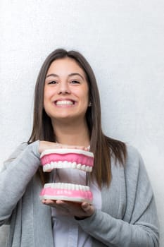 Close up portrait of attractive young girl holding oversize human teeth prosthesis.Woman  with fun facial expression against seamless light background.