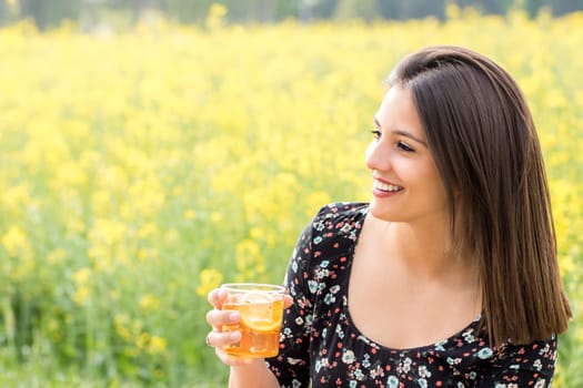 Close up portrait of attractive young woman holding glass with ice tea in yellow flower field.