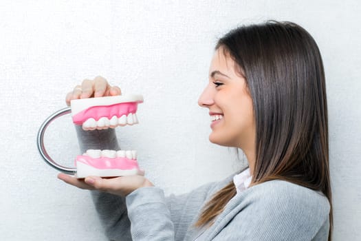 Close up portrait of attractive young girl holding oversize human teeth prosthesis.Side view of woman against light textured background.