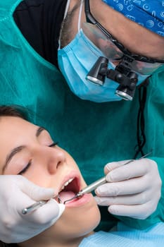 Close up of dentist wearing medical magnifying glasses working on female patient.Surgeon in green gown with mouth mirror and scaler.