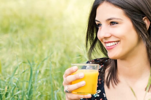 Close up face shot of cute young woman with charming smile drinking orange juice outdoors.