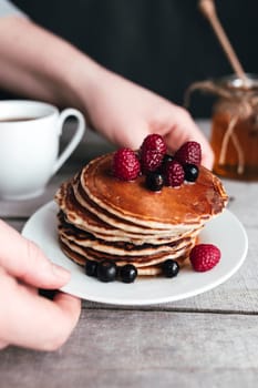 Hands hold white plate with pancakes, berries, honey, coffee cup on wooden table, jar and spoon. High quality photo