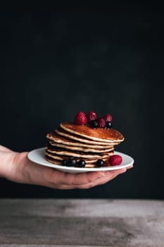 Juicy pancakes with berries and honey on white plate on human hand, wooden table. High quality photo