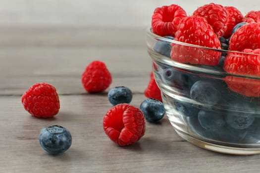Close up, glass bowl full of blueberries with raspberries on top, some of them spilled on gray wood desk.