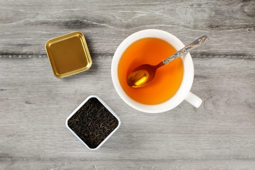 Table top view on cup of hot amber tea with spoon, and caddy full of black dried loose leaves, placed on gray wood desk.