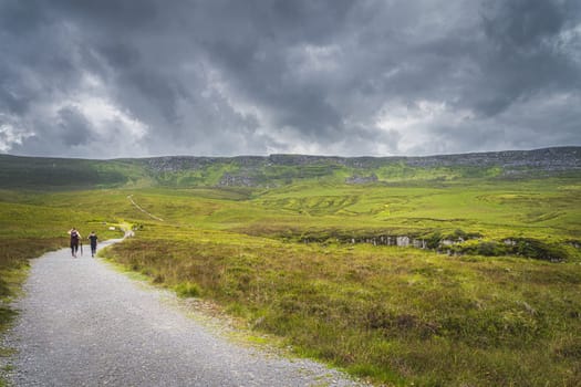People hiking on a trail between green hills and peat bog leading to Cuilcagh Mountain with stormy, dramatic sky in background, Northern Ireland