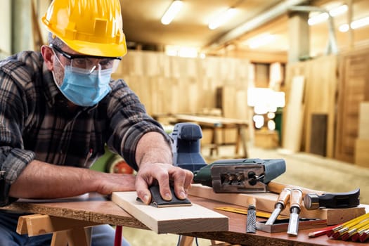 Carpenter worker at work in the carpentry workshop, wears helmet, goggles and surgical mask to prevent coronavirus infection. Preventing Pandemic Covid-19 at the workplace.