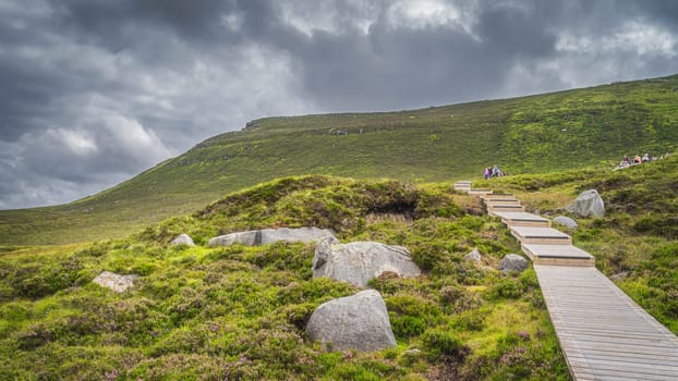 People hiking on wooden boardwalk, between boulders and heathers, leading to Cuilcagh Mountain. Storm, dramatic sky in background, Northern Ireland
