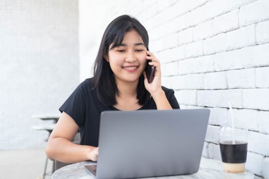 Asian woman freelance talking on mobile phone while typing on laptop in coffee shop. Young female with cute smile having talking with cellphone during work on portable laptop computer.