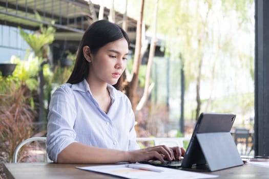 Young Asian woman freelance is working with tablet at coffee shop. Business woman typing, searching and checking document in modern cafe.  Working online technology system concept.