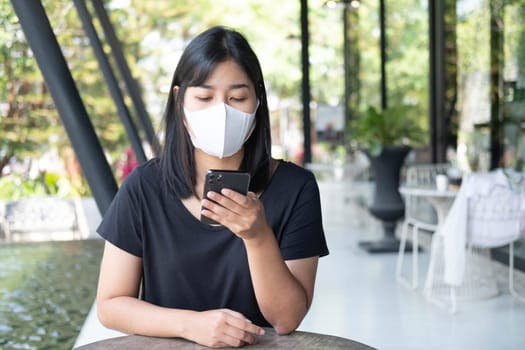 Young Asian woman wearing medical mask using smartphone at coffee shop. Asian female reading text message or social media on cell telephone during in modern cafe. New normal and social distance working.