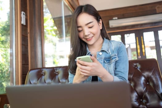Smiling young Asian woman using smartphone at coffee shop. Asian female reading text message or social media on cell telephone during in modern cafe. Working online technology system concept.