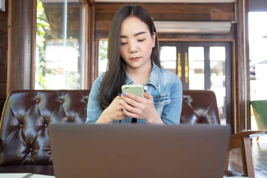 Smiling young Asian woman using smartphone at coffee shop. Asian female reading text message or social media on cell telephone during in modern cafe. Working online technology system concept.