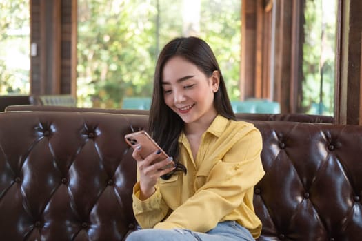 Smiling young Asian woman using smartphone at coffee shop. Asian female reading text message or social media on cell telephone during in modern cafe. Working online technology system concept.