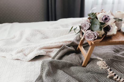Romantic morning. Wooden coffee table with flowers on bed with plaid, coffee cup, flowers and candles. Lilac roses with eucalyptus and anemones. Interior gray tones. High quality photo