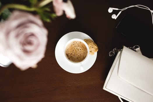 A cup of coffee with a bouquet of tender anemone, eucalyptus branches and pastel purple roses, a woman handbag, headphones and a phone on a brown wooden table. Blurred background