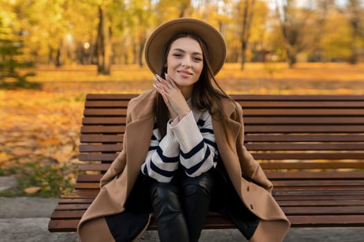 portrait of attractive stylish smiling woman with long hair walking in the park, dressed in warm brown coat autumn trendy fashion, street style wearing hat.