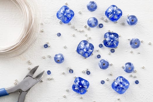 Blue and white glass beads with silver sead beads, wire cutters, and beading wire, on white painted wooden surface, top down view