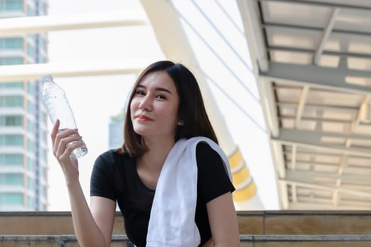 Sportive girl with slim body in black sportswear and white towel over her shoulder sitting down on stairs in modern city, holding mineral water bottle and taking a breake from athlete jogging outdoors or work out